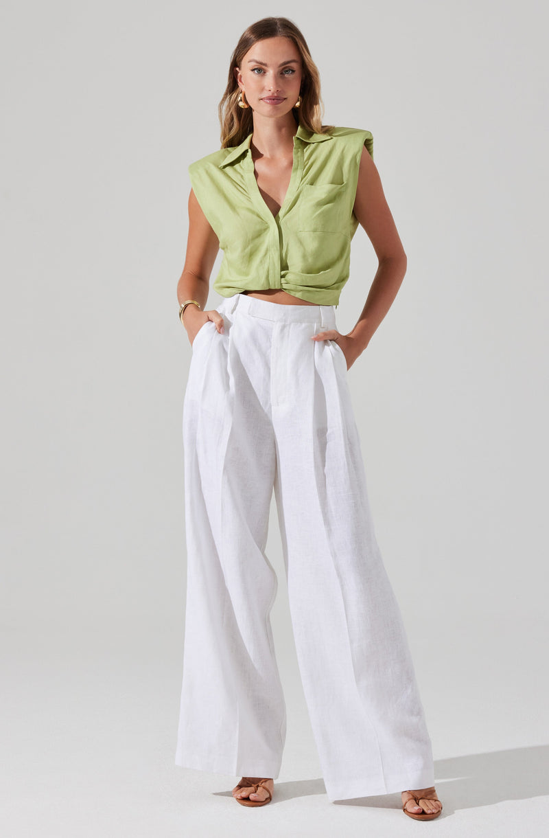 A woman is wearing a sleeveless, light green shirt with a collar and a front pocket, styled in a relaxed, slightly cropped fit. She pairs it with high-waisted, wide-leg white linen pants that feature pleats at the waist and side pockets. She accessorizes with gold earrings, a gold bracelet, and tan open-toe heels.