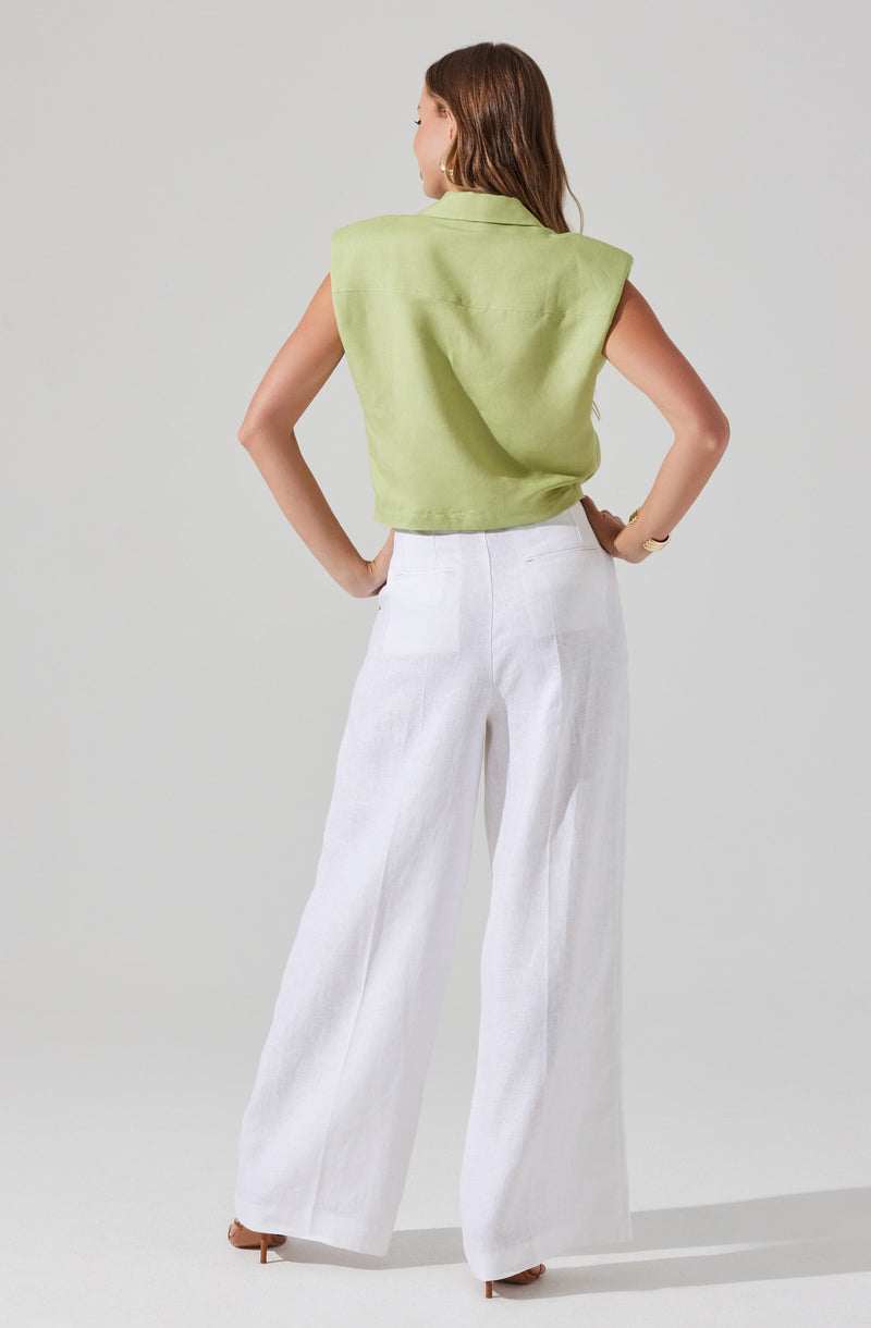 A woman is wearing a sleeveless, light green top with broad shoulder accents and a collar. She has paired it with high-waisted, wide-leg white pants and brown heeled shoes.