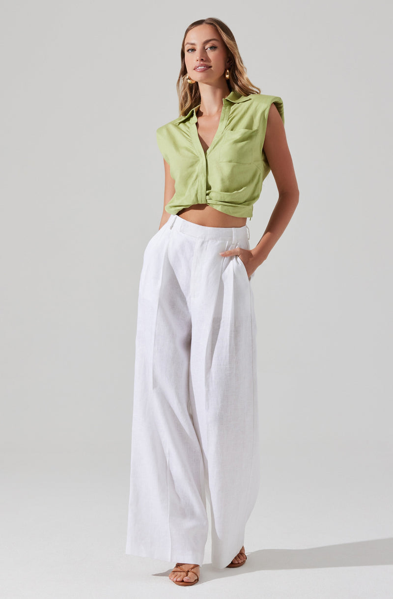 A woman is wearing a sleeveless, lime green, collared button-up top that is tied at the waist, paired with high-waisted, wide-leg white pants.
