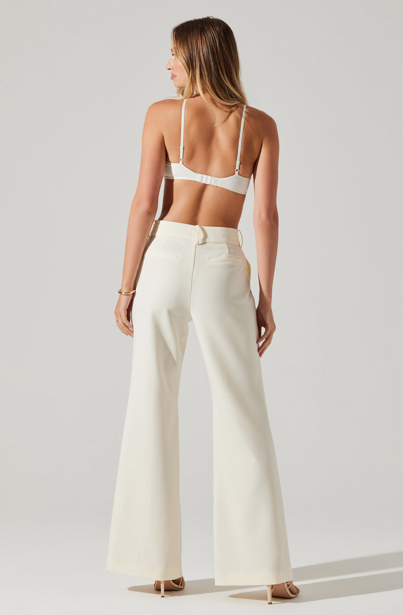 A woman is wearing high-waisted, wide-leg white trousers with belt loops and a back zipper. She is also wearing a white bra with adjustable shoulder straps. The overall look is elegant and minimalistic.
