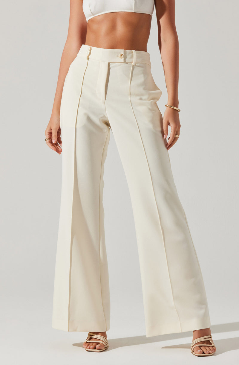 A woman is wearing high-waisted, wide-leg cream-colored pants with visible vertical seam details. She is also wearing a white cropped top that exposes her midriff, and tan strappy high-heeled sandals. She has a bracelet on her right wrist.