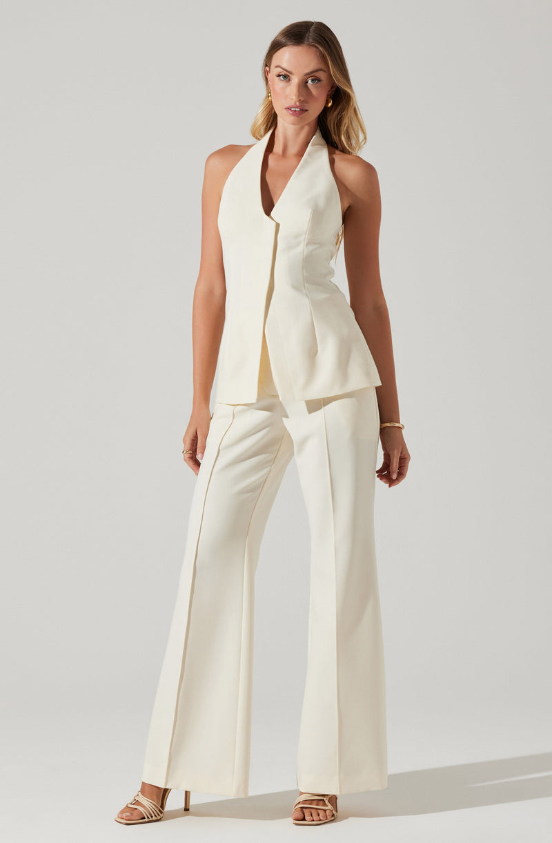 A woman is wearing an elegant, sleeveless cream-colored pantsuit. The top features a high collar and a deep V neckline, with a tailored, structured fit that flares slightly at the hem. The matching wide-leg trousers have a relaxed fit and a subtle crease down the front. She is also wearing nude strappy high-heeled sandals and simple gold jewelry.