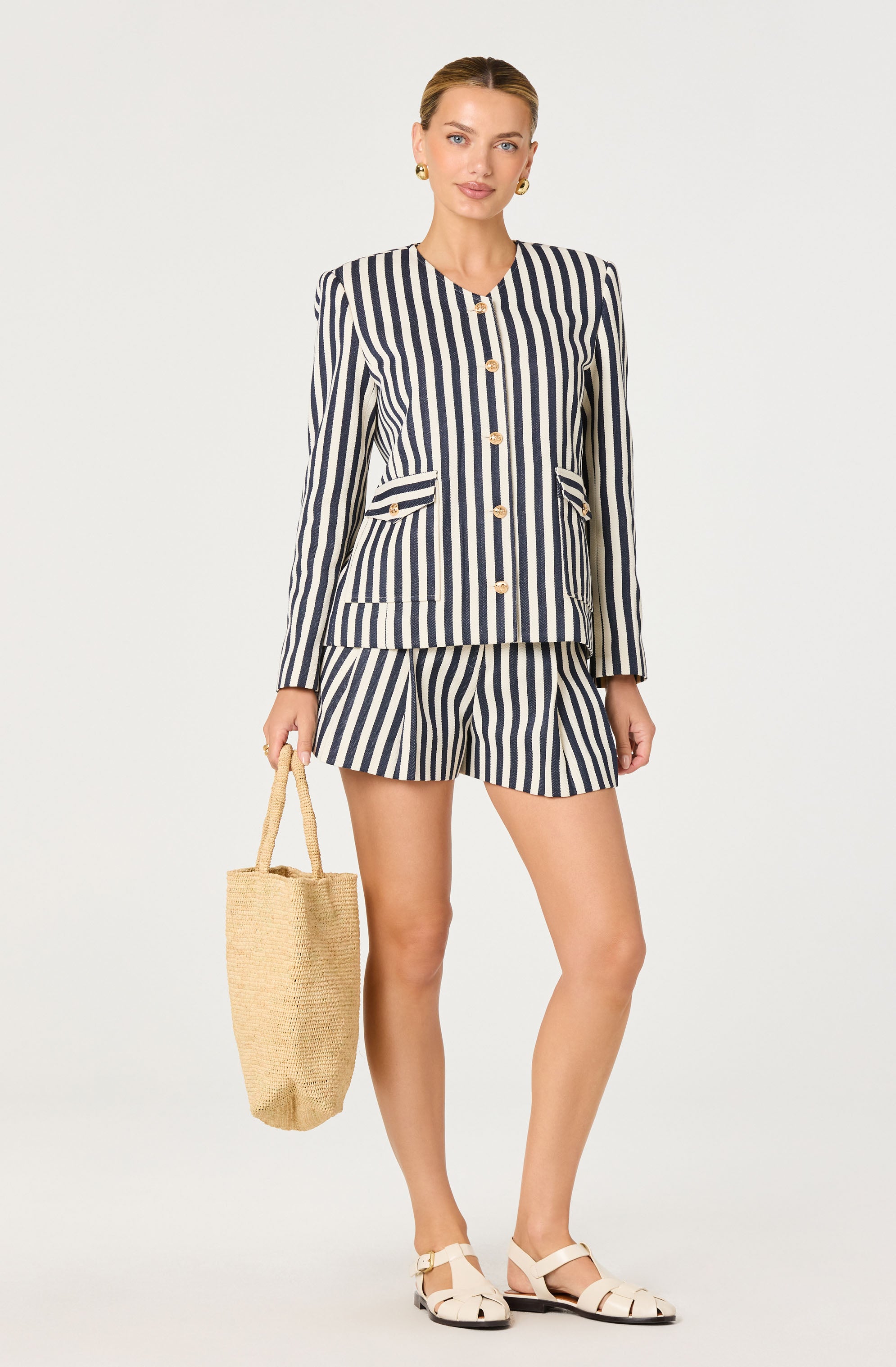 Woman in a navy and white striped blazer with gold buttons and matching shorts, holding a straw bag.