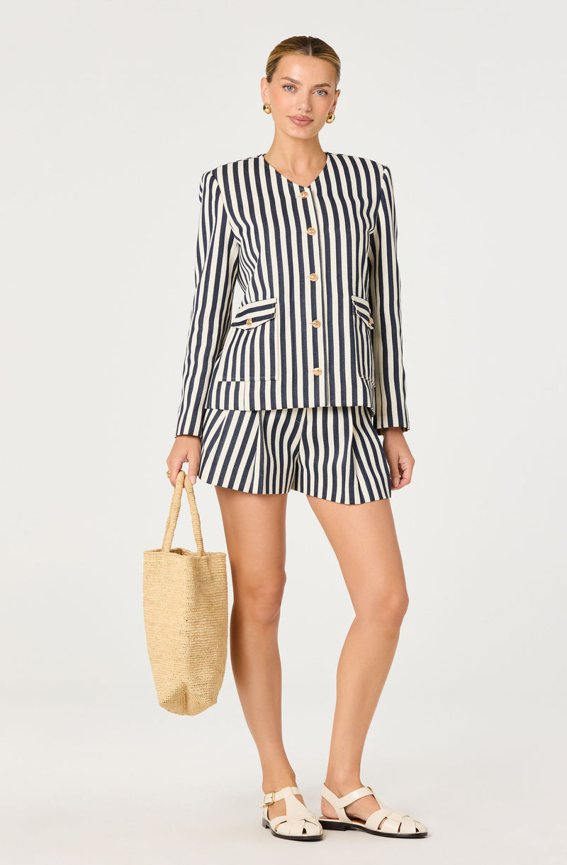 Woman in a navy and white striped blazer with gold buttons and matching shorts, holding a straw bag.