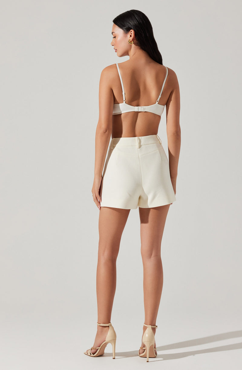 A woman is wearing a white two-piece outfit consisting of a fitted crop top with thin straps and a back clasp, paired with high-waisted, tailored shorts. She is also wearing beige high-heeled sandals with ankle straps.
