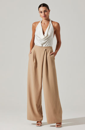 A woman is wearing a sleeveless white draped top with a cowl neckline, paired with high-waisted, wide-leg beige trousers that have a single button closure and side pockets. She is also wearing white open-toe heels and gold hoop earrings.
