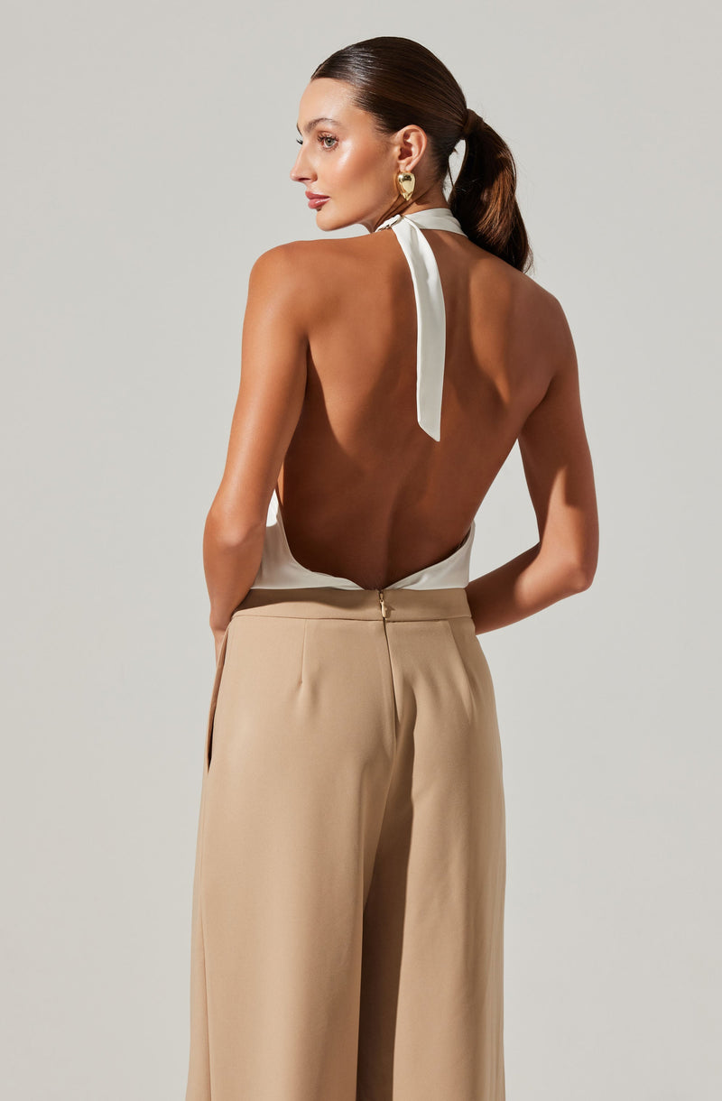 A woman is wearing a sleeveless, backless white halter top with a tied bow at the neck. She pairs it with high-waisted, beige wide-leg trousers. She accessorizes with large gold hoop earrings.