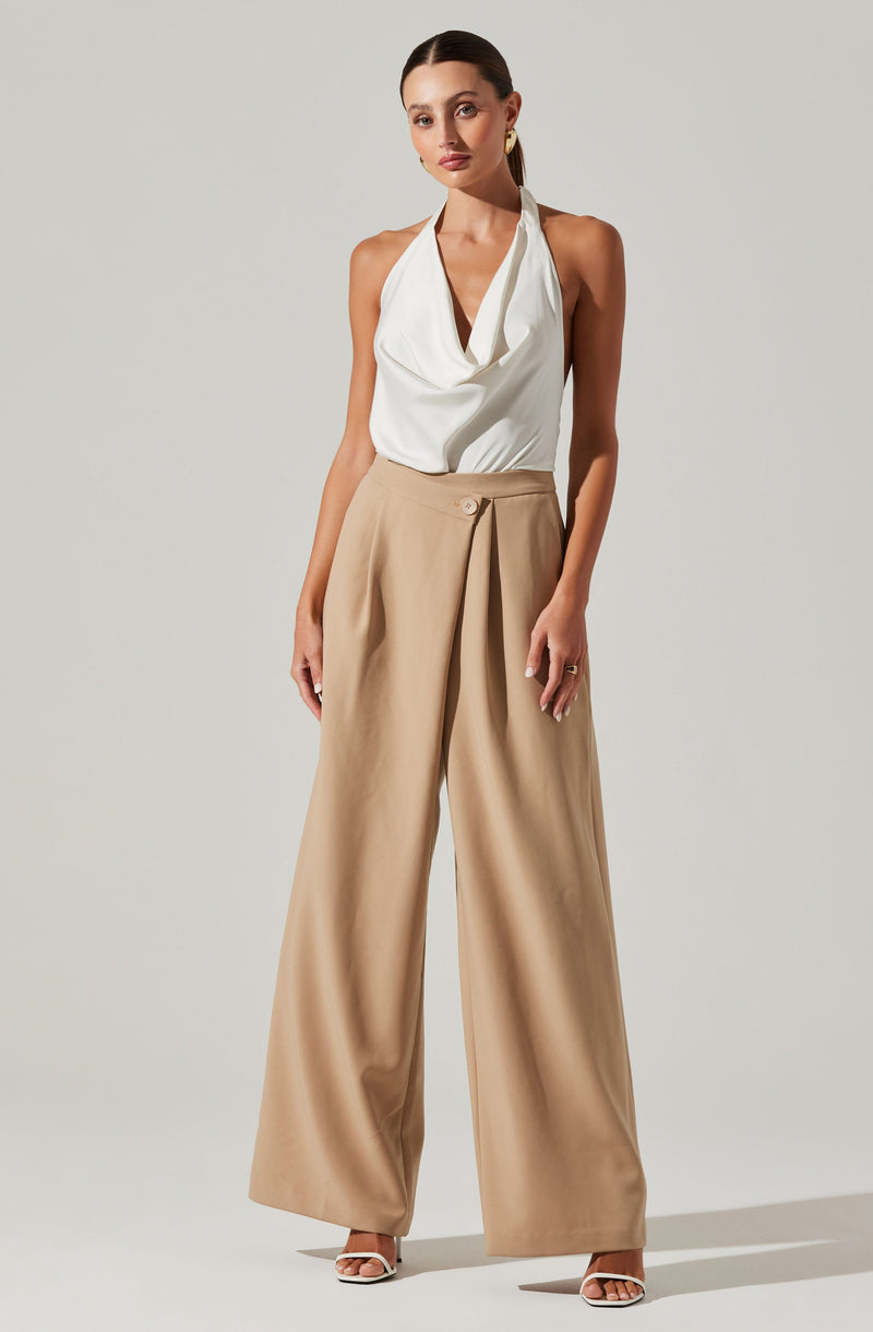 A woman is wearing a sleeveless, white halter top with a draped neckline and beige high-waisted, wide-leg trousers. She has accessorized with gold earrings and a ring on her right hand, and is wearing open-toe heels.