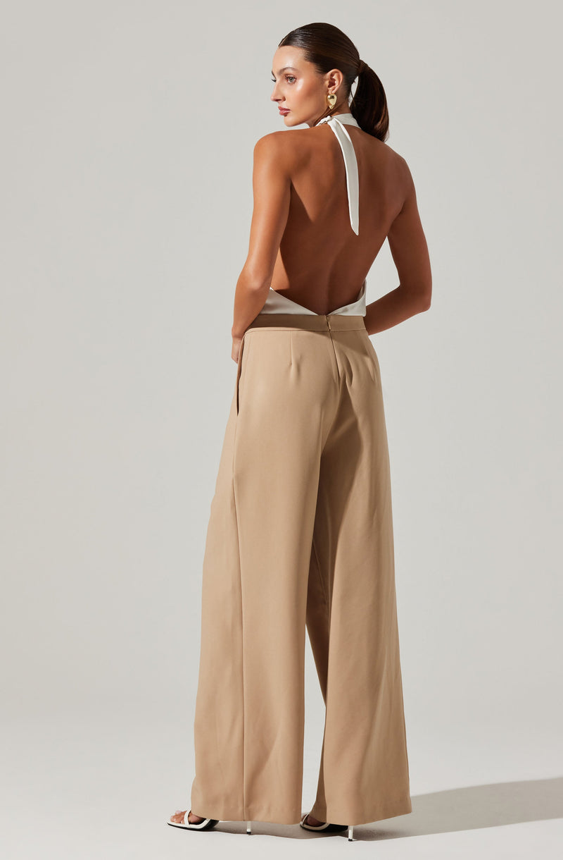 A woman is wearing high-waisted, wide-leg beige trousers paired with a sleeveless, backless top that ties around the neck with a white strap. She is accessorized with large gold earrings and stands against a plain, light-colored background.