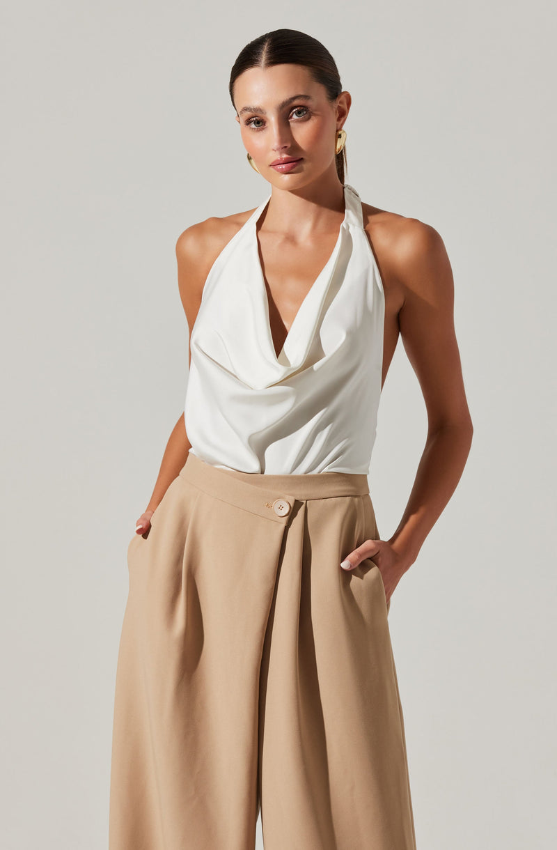 A woman is wearing a sleeveless white top with a draped neckline and beige high-waisted wide-leg trousers with pleats and a button closure.