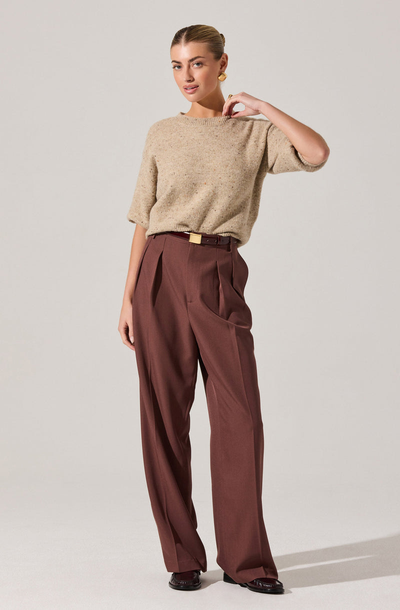 A woman is wearing a beige, loose-knit sweater with a round neckline and three-quarter length sleeves. She pairs it with high-waisted, wide-leg brown trousers that have pleats at the waist and a black belt with a gold buckle. She completes her look with dark shoes and a large gold earring.