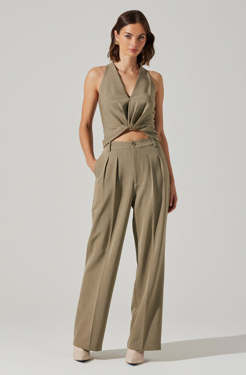 The woman is wearing a sleeveless, olive-green top with a deep V-neckline, which is tied in a knot at the front, and matching high-waisted, wide-leg trousers with pleats at the waist. She has on light-colored pointed-toe shoes and is accessorized with earrings.