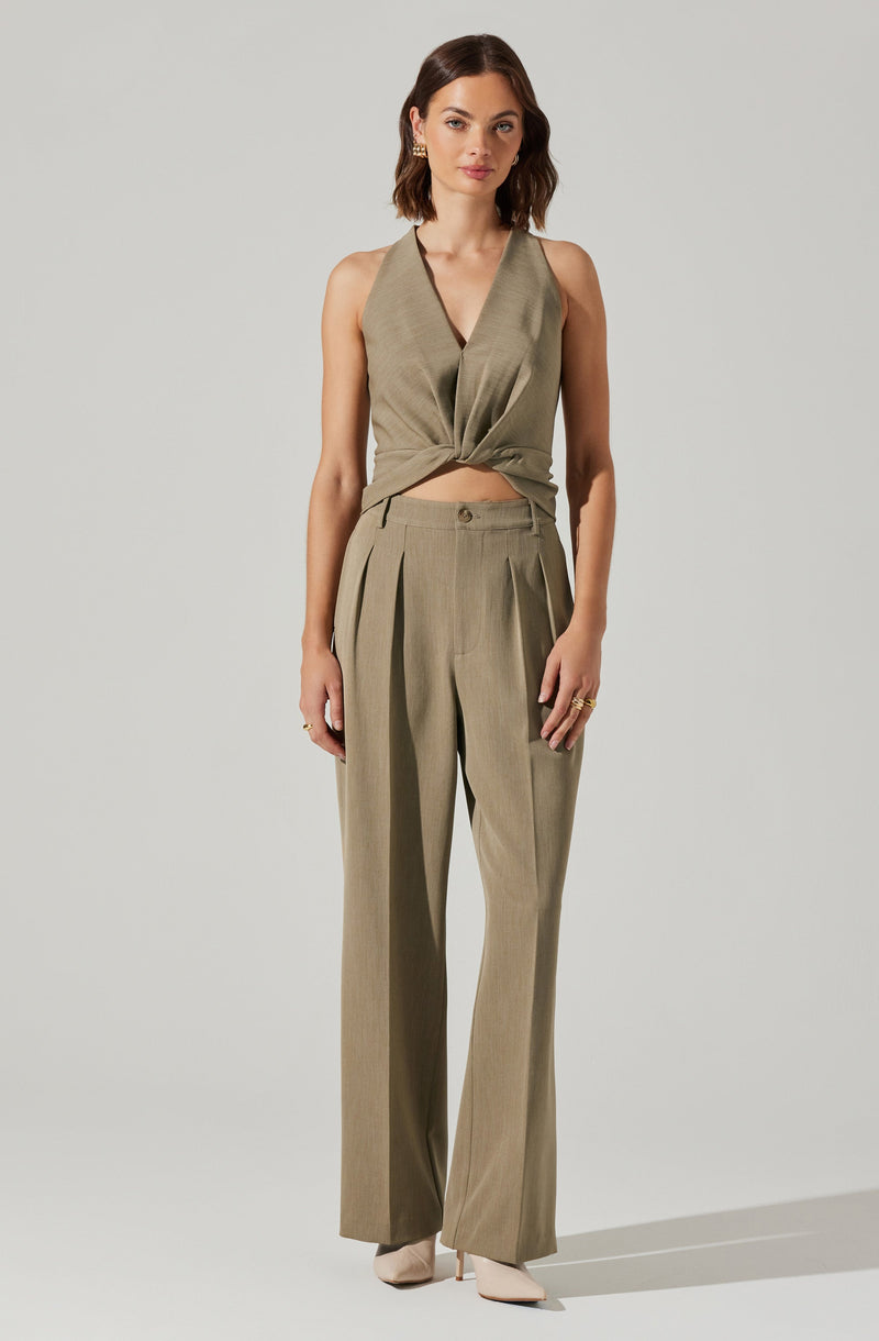 A woman is wearing a sleeveless, taupe-colored top with a deep V neckline that is knotted at the waist, pairing it with matching wide-leg trousers featuring pleats at the waist. She has on pointed-toe shoes in a light color and accessorizes with gold earrings and rings.