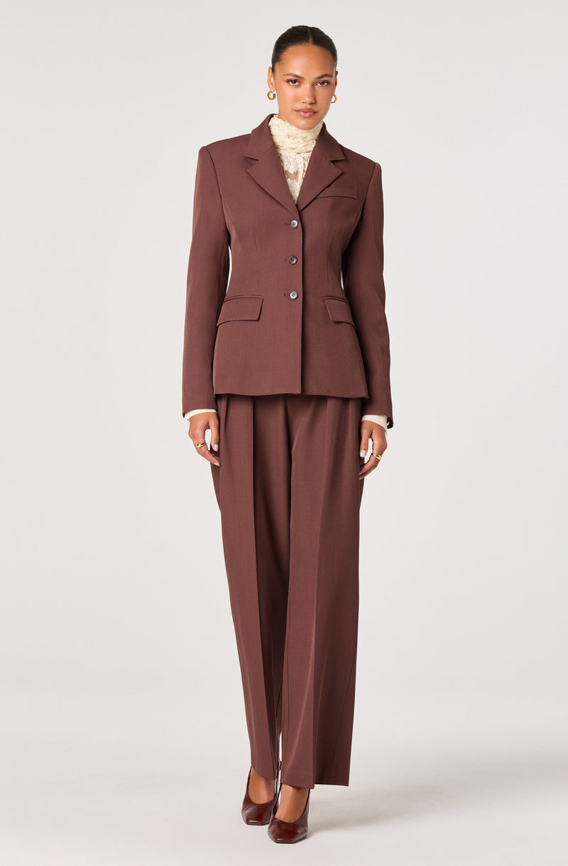 A woman is wearing a fitted, brown tailored blazer with notch lapels, three buttons down the front, and two flap pockets at the waist. She has on matching wide-leg brown trousers with a high waist and pleats. Under the blazer, she is wearing a cream-colored, textured turtleneck sweater. She completes the look with dark brown pointed-toe heels and gold earrings.
