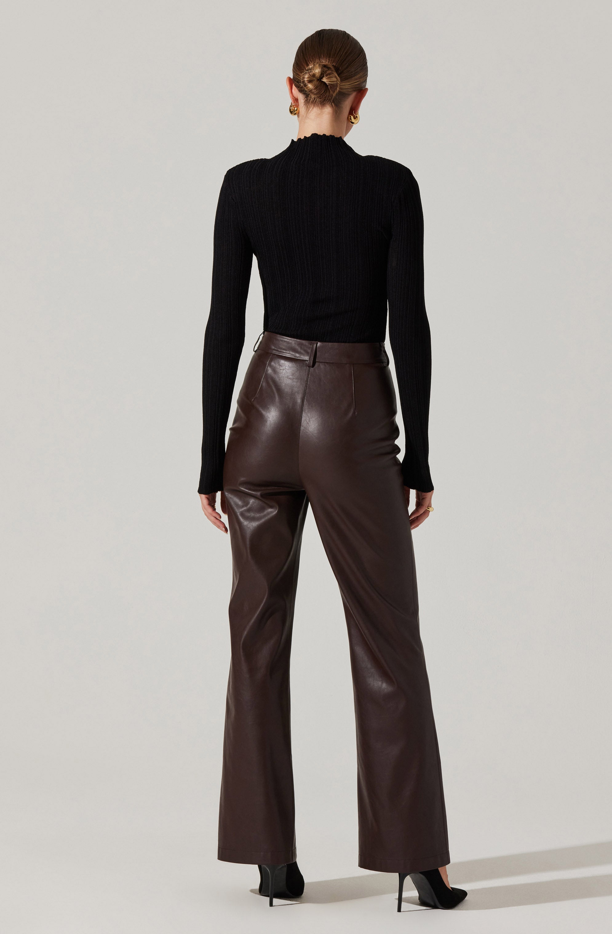 A person is wearing a fitted, long-sleeve, ribbed black sweater with a high neckline. They are also wearing high-waisted, dark brown leather pants with a straight-leg cut, paired with black high-heeled shoes.