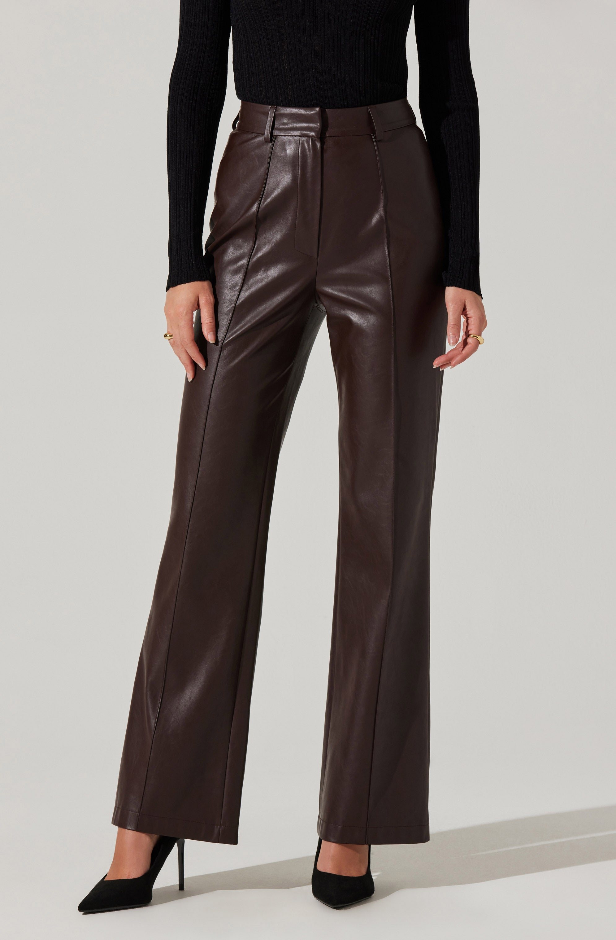 A person is wearing high-waisted, wide-leg brown leather pants with a front zipper and belt loops. They are paired with a black long-sleeve, ribbed sweater and black high-heeled shoes. The individual has minimal jewelry, including rings on both hands.