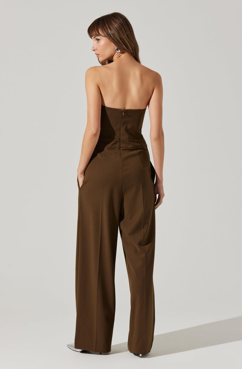 A woman is wearing a strapless, brown jumpsuit with wide-leg pants. The fabric appears smooth and slightly shiny. The back of the jumpsuit features a concealed zipper running down the center. The woman has her hands in her pockets and is standing against a plain, light gray background.