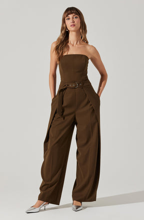 A woman is wearing an olive-green strapless jumpsuit with a fitted bodice and wide-leg pants. The jumpsuit features a matching fabric belt with grommet details at the waist, and the pants have pleats at the hips. She is also wearing pointed-toe silver heels.