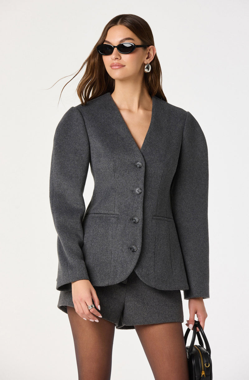A woman is wearing a tailored, dark gray blazer with a V-neckline and four buttons down the front. She has on matching gray shorts, sheer black tights, and accessorizes with large hoop earrings, black sunglasses, multiple rings, and is holding a small black handbag.