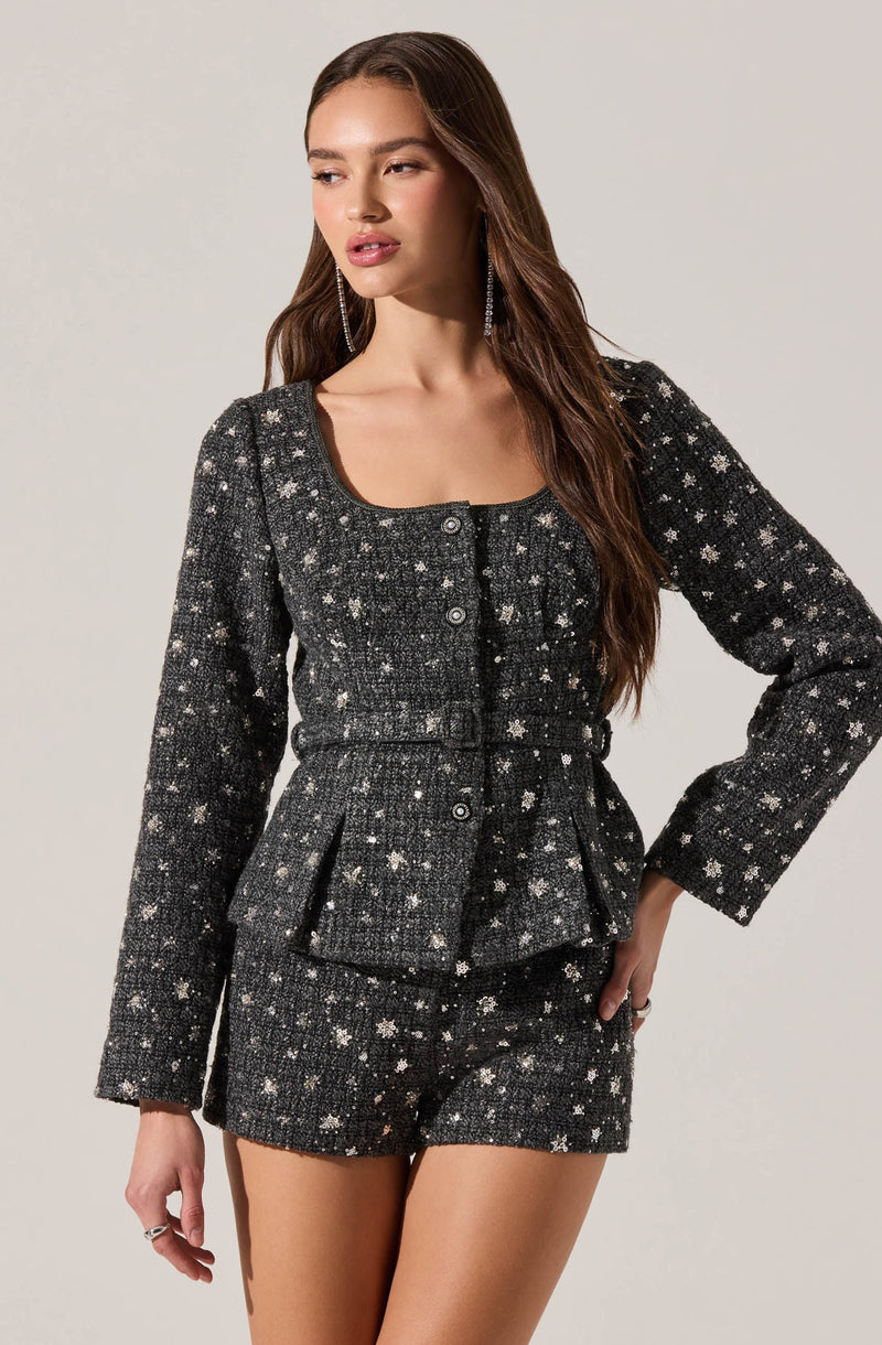 A woman is wearing a matching black tweed suit with a textured pattern of white and silver stars. The suit includes a fitted, long-sleeved jacket with a rounded neckline, button-front closure, and two flap pockets at the waist. She is also wearing high-waisted shorts made from the same star-patterned tweed fabric.