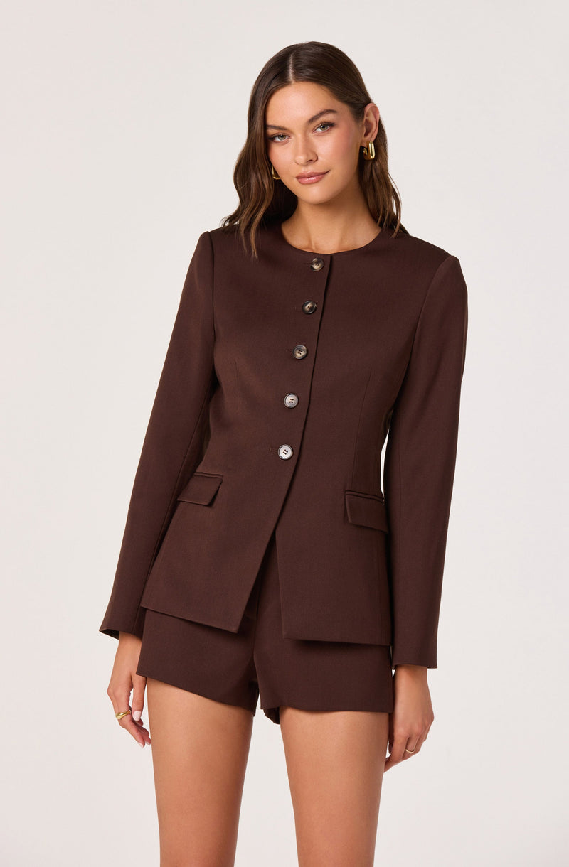 A woman is wearing a tailored, dark brown blazer with a round neckline, featuring a row of large buttons down the front. The blazer has long sleeves and two flap pockets on the front. She pairs it with matching dark brown shorts.