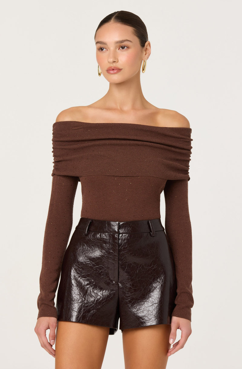 A woman is wearing a brown off-the-shoulder sweater with a wide neckline and long sleeves. The sweater appears to be fitted and slightly textured. She pairs it with high-waisted, shiny black leather shorts.