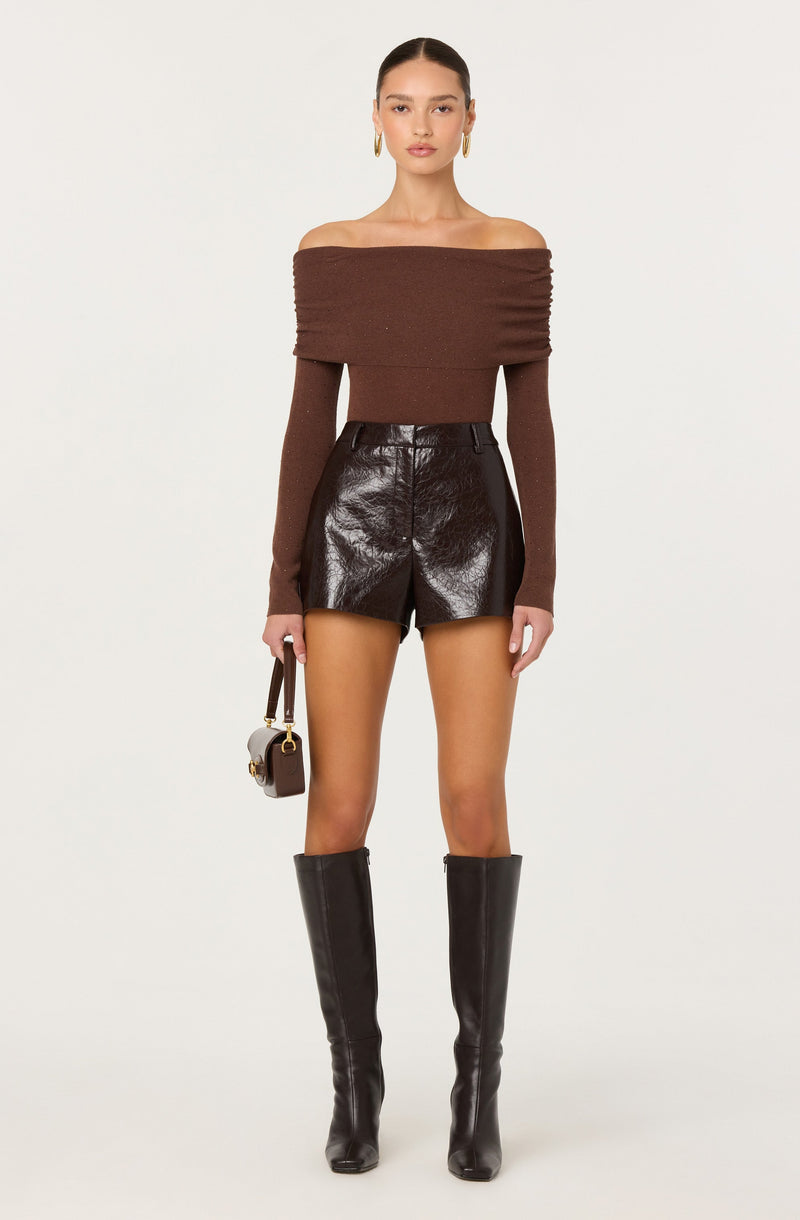A woman is wearing a brown off-the-shoulder sweater with long sleeves, paired with high-waisted, shiny black leather shorts. She accessorizes with gold hoop earrings, carries a small handbag with a strap, and wears tall black leather boots that reach just below the knee.