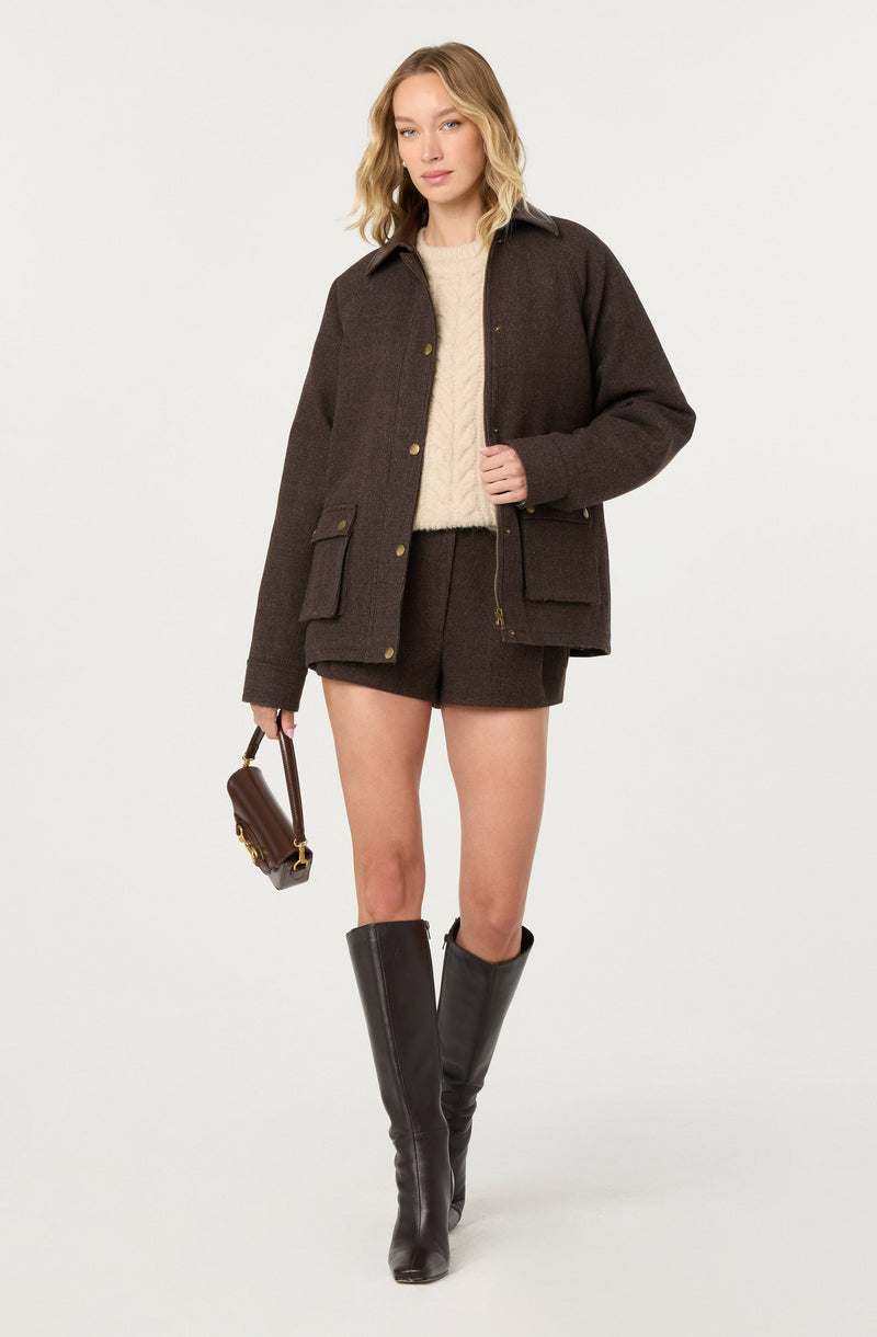 A woman is wearing a dark brown wool jacket with large front pockets, a cream-colored cable-knit sweater underneath, matching dark brown shorts, and tall black leather knee-high boots. She is holding a small dark brown handbag with a gold clasp and chain strap.