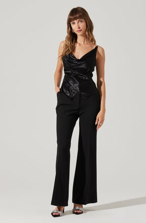 A woman is wearing a sleeveless black satin top with a draped, cowl neckline and asymmetrical hem, paired with high-waisted black wide-leg trousers. She is also wearing open-toed high heels with sparkly embellishments.