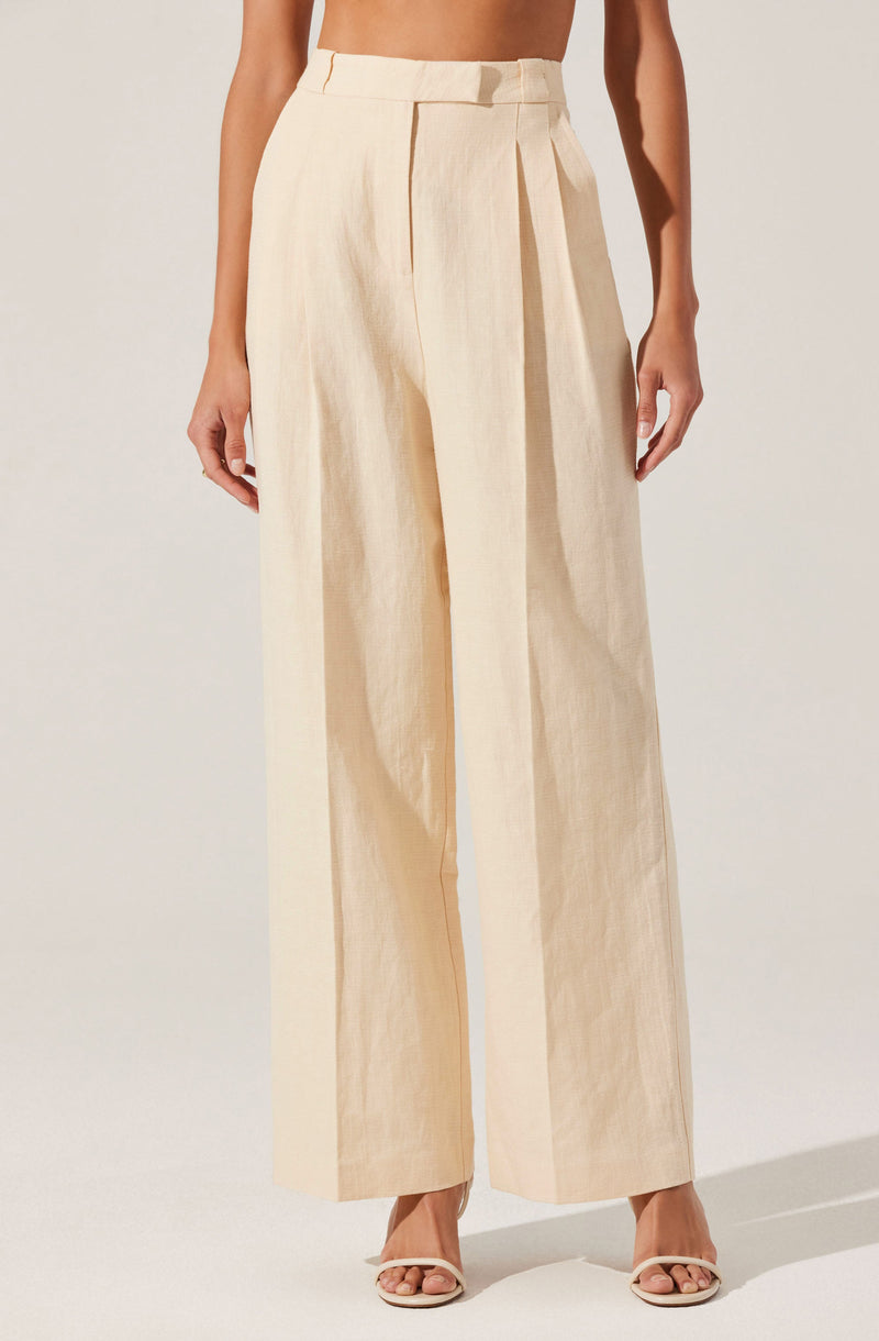 A person is wearing high-waisted, wide-leg trousers in a light beige or cream color. The trousers feature a belt loop detail at the waistband and a prominent front pleat that runs down each leg. The fabric appears to be lightweight and slightly textured. The outfit is paired with open-toed heels. The person's arms are relaxed by their sides, and the background is plain and neutral.