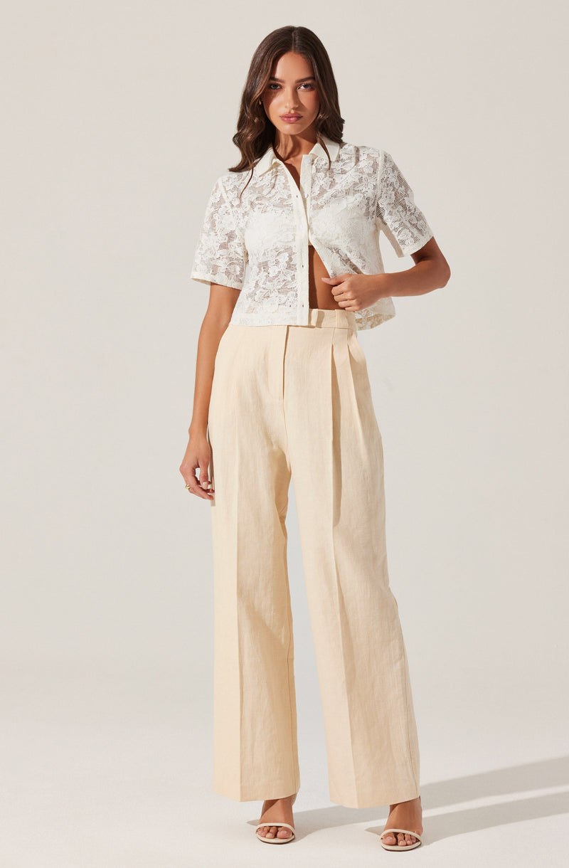 A woman is wearing a short-sleeved, white lace cropped jacket with a collar and front zipper, paired with high-waisted, wide-leg cream trousers. She is also wearing beige open-toed heels.