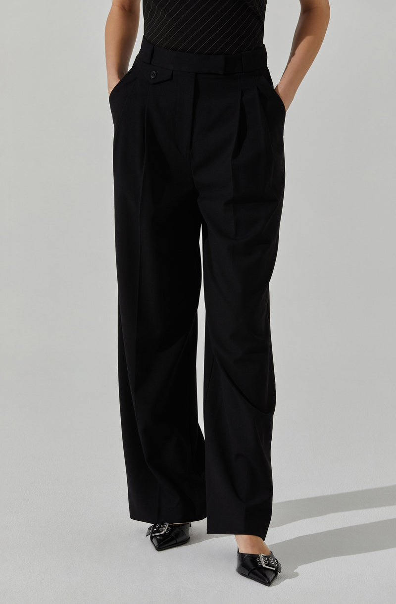 A person is wearing high-waisted, wide-leg black trousers with pleats at the waist, featuring belt loops and a button closure at the front. They are also wearing a black top with thin, diagonal pinstripes. The person's hands are in their pockets. They are wearing black pointed-toe shoes with lace-up details and small metallic accents.
