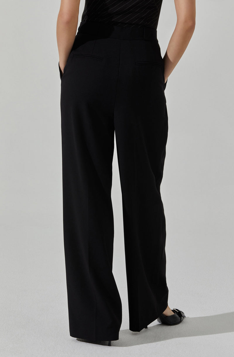The person is wearing high-waisted black dress pants with a wide-leg fit and back welt pockets. They are also wearing black shoes with a small bow detail.
