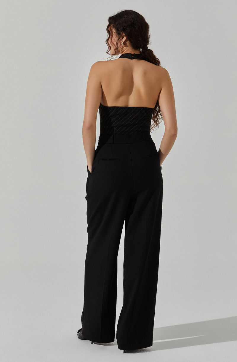 A woman is wearing a strapless black top with a subtle linear pattern and a high neckline. She pairs it with high-waisted, wide-leg black trousers that have a tailored fit and belt loops. Her hair is long, curly, and dark, styled loosely over her shoulders.