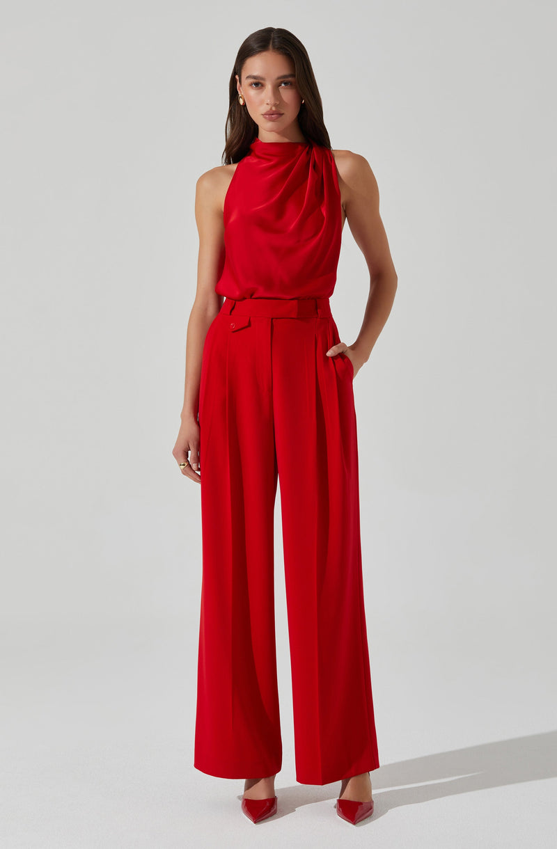 A woman is wearing a sleeveless, high-neck red blouse with draped fabric at the neckline, paired with high-waisted, wide-leg red trousers with pleats and a matching belt. She completes the outfit with red pointed-toe shoes.