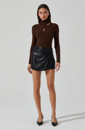 A woman is wearing a fitted, long-sleeve, brown top with a high neckline and cut-out details near the collarbone and shoulders. She pairs it with a black leather mini skirt featuring a gathered, ruched design on one side. She completes the outfit with black pointed high heels that have a small bow detail.