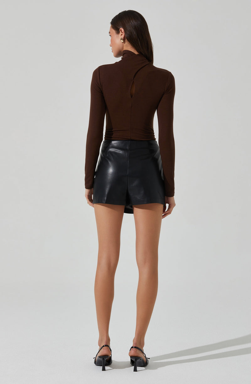 A woman is wearing a fitted, long-sleeved, high-neck brown top with a small keyhole opening at the back. She pairs it with a high-waisted black leather mini skirt that has a center back slit. She completes the outfit with black open-toe high-heeled sandals and gold earrings.