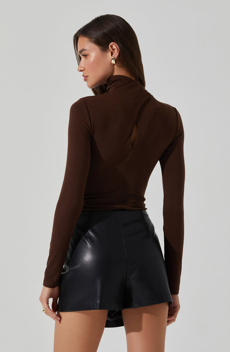 A woman is wearing a fitted, long-sleeved, brown turtleneck top with a small keyhole cutout at the back. She pairs it with high-waisted black leather shorts.