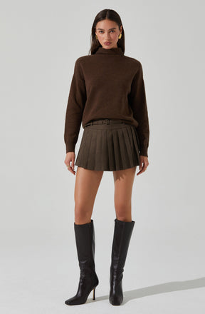 A woman is wearing a brown high-neck sweater with long sleeves, a belt, a plaid pleated skirt with a mix of brown and gray tones, and black knee-high heeled boots. She has long dark hair and is accessorized with large gold earrings.