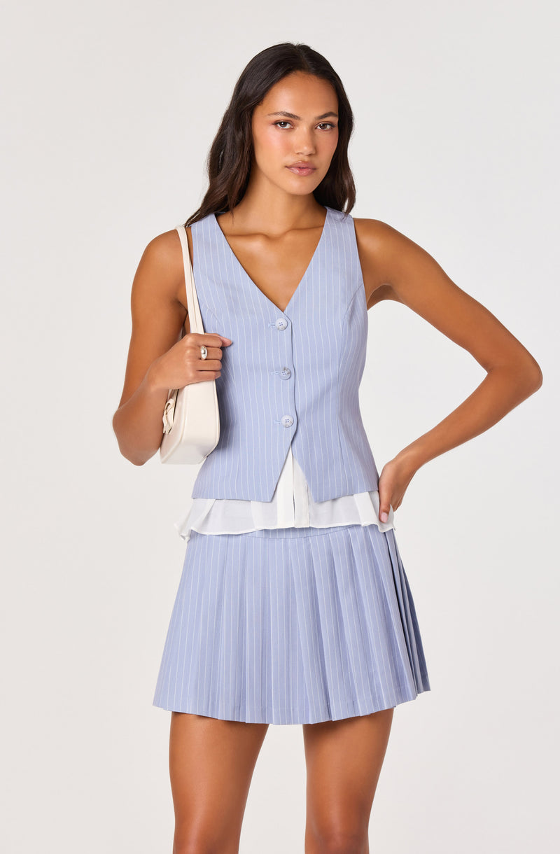 Woman in a light blue pinstripe vest and pleated skirt, holding a white bag over her shoulder.