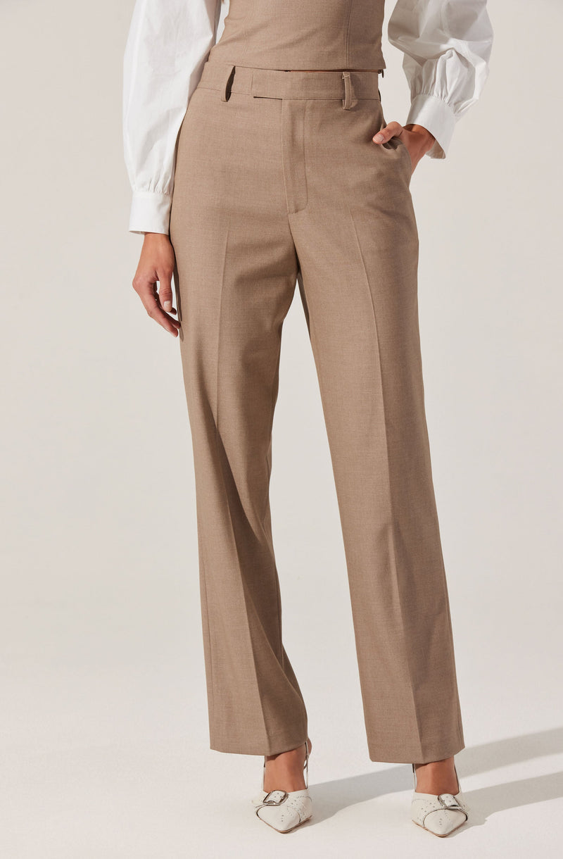 A person is dressed in beige dress pants with a high waist and pleats at the front. The pants have belt loops but no belt is worn. They are paired with a white blouse with long, slightly puffed sleeves and wide cuffs. The person is also wearing white pointed-toe shoes with a small heel.