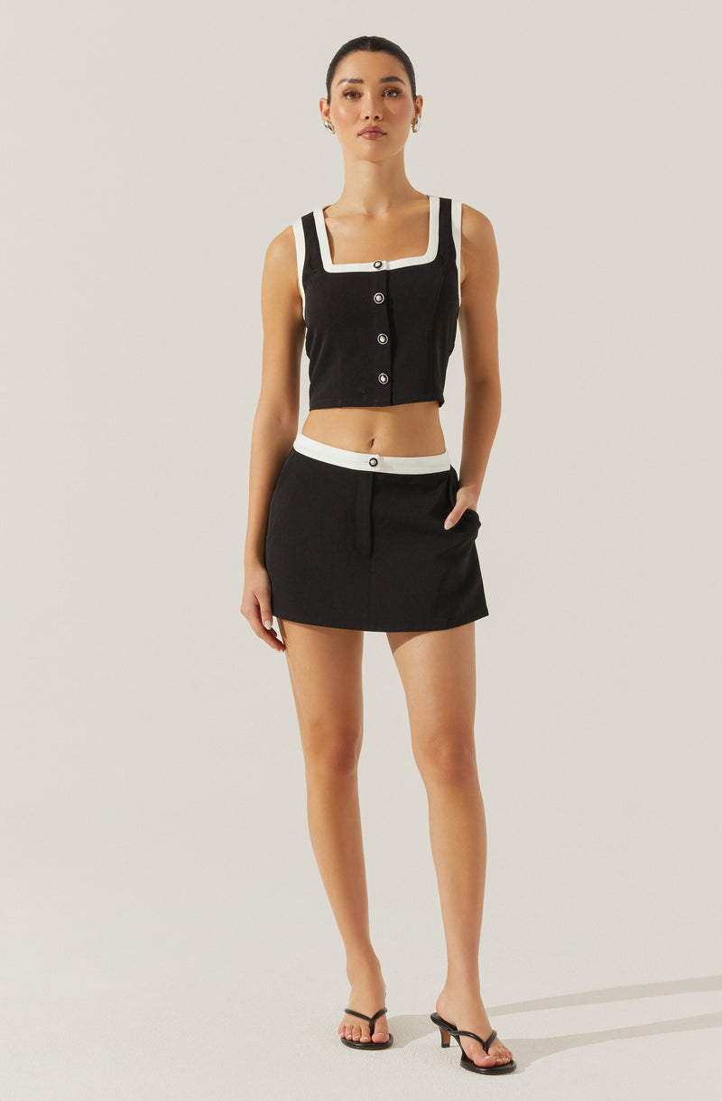 A woman is wearing a sleeveless, cropped black top with white accents around the square neckline, shoulder straps, and button placket. She is also wearing a high-waisted black mini skirt with a white waistband and a front button. She has on black high-heeled sandals and small hoop earrings.