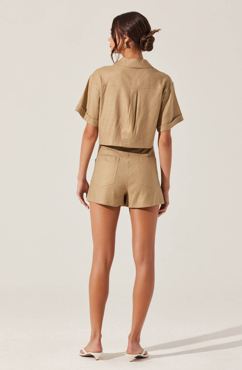 A woman is wearing a matching beige short-sleeved shirt and high-waisted shorts set. The shirt has a loose fit, a collar, and rolled-up sleeves. The shorts have a relaxed fit with visible back pockets. She is also wearing white heeled sandals.