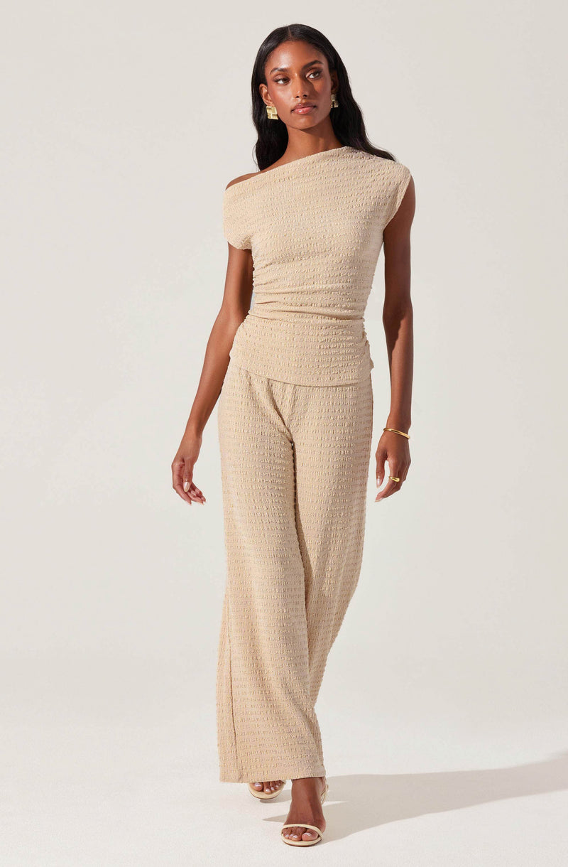 A woman is wearing a matching beige outfit consisting of a textured sleeveless top with a one-shoulder design and wide-leg pants with a similar textured pattern. She has long, dark hair and is accessorized with gold earrings, a gold bracelet, and rings. She is standing against a plain, light background.