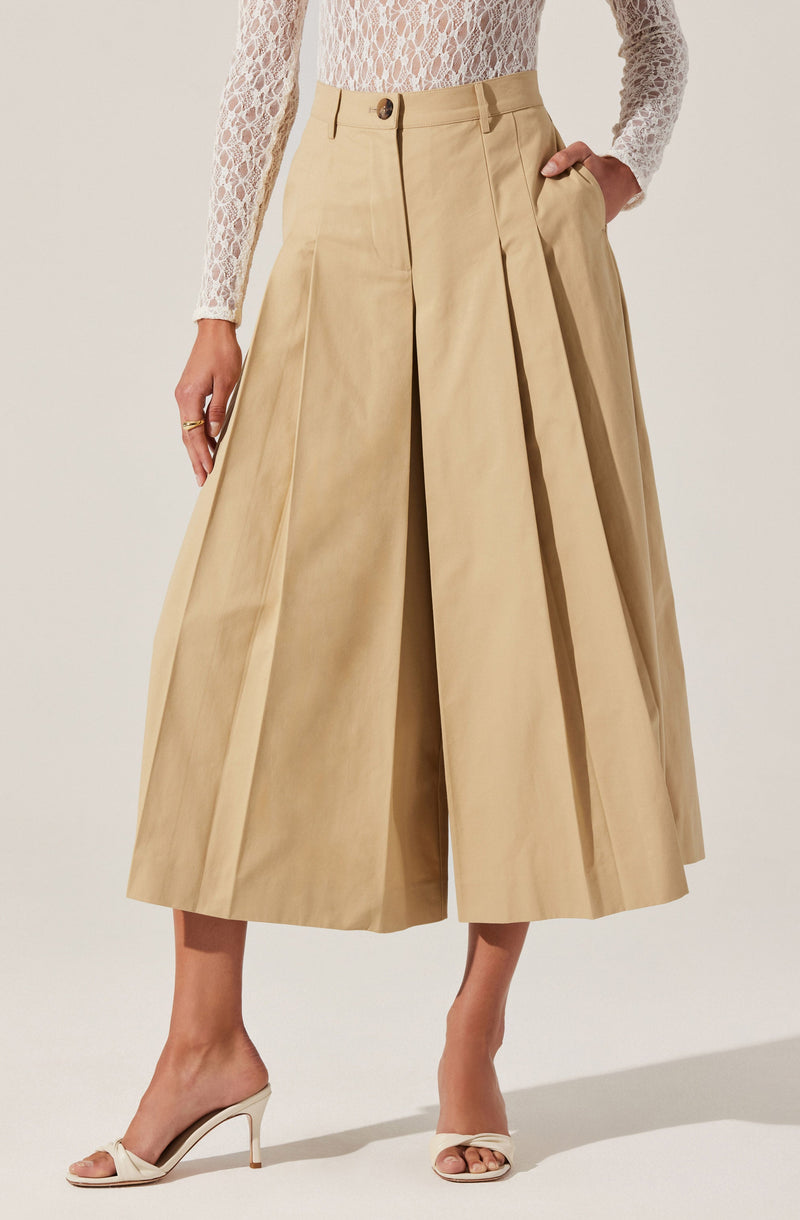 A person is wearing high-waisted, wide-leg beige pants with pleats at the front. The pants have a single button and zipper closure, and belt loops at the waistband. The individual is also wearing a white, long-sleeved, lace top with a floral pattern. They have on open-toed, beige high-heeled sandals with a bow detail on the front.