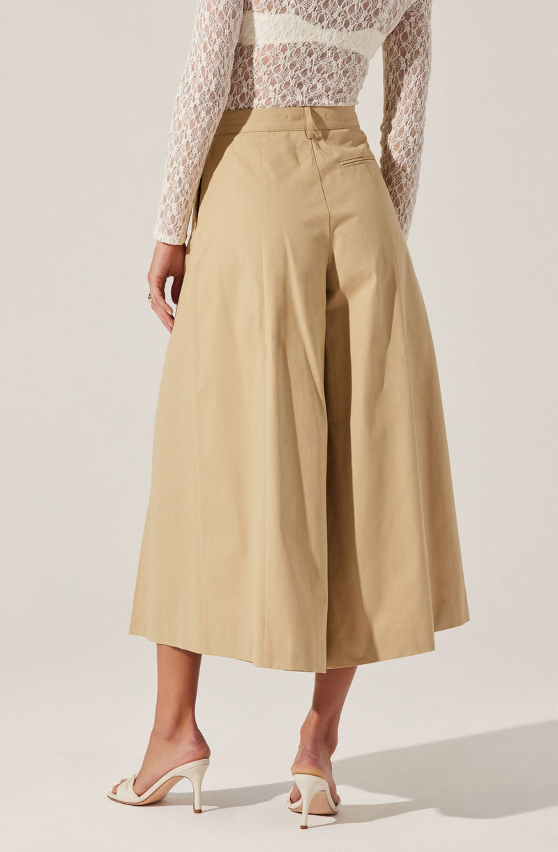 A woman is wearing high-waisted, wide-leg beige culottes that reach mid-calf. She pairs them with a white, long-sleeved lace top. She completes the look with white high-heeled sandals featuring a bow detail on the front strap.