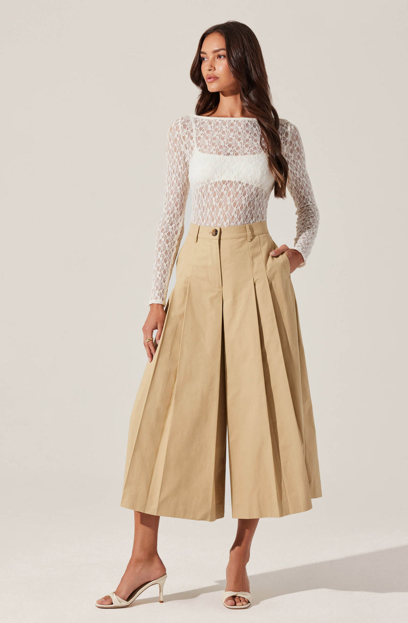 A woman is wearing a long-sleeved, see-through white lace top with a floral pattern. She has paired it with high-waisted, wide-leg beige pants that have front pleats and side pockets. She is also wearing open-toe, ankle-strap high-heeled sandals in a light color.