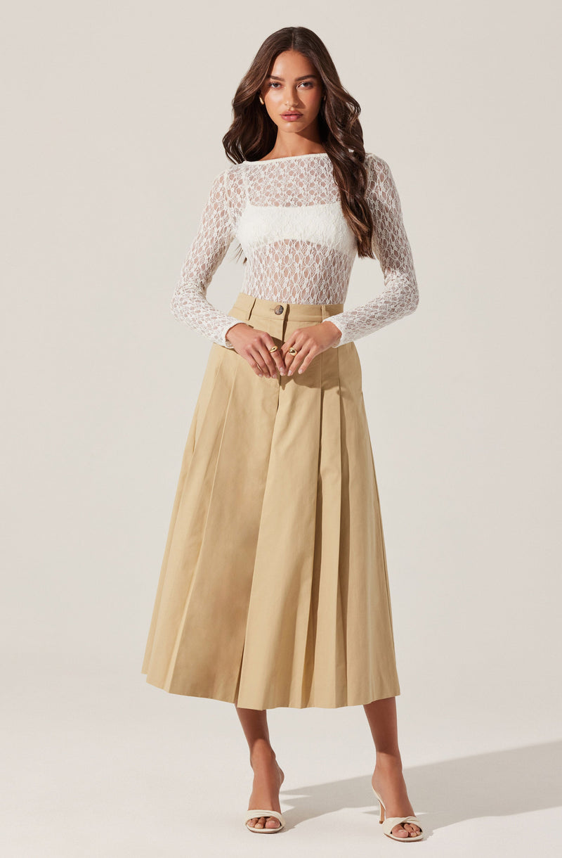 A woman is wearing a long, cream-colored lace top with a square neckline and long sleeves. She pairs it with a high-waisted, beige pleated midi skirt and white open-toe heels.