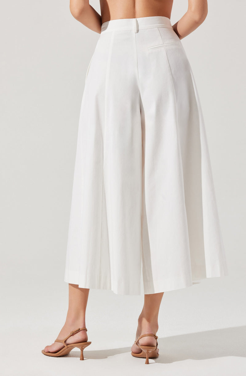 A person is wearing high-waisted, wide-leg white pants that reach mid-calf. The pants have a smooth front with no visible buttons or zippers, and feature a back pocket on the right side. The model is also wearing nude-colored, open-toe heeled sandals.