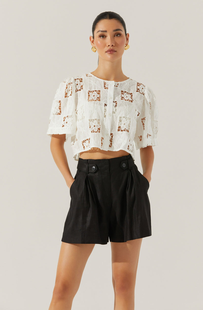 A woman is wearing a white, short-sleeved, lace top with a floral and geometric pattern. The top is cropped, ending above her waist. She pairs it with high-waisted black shorts that have pleats, a button detail at the waistband, and a slightly loose fit.